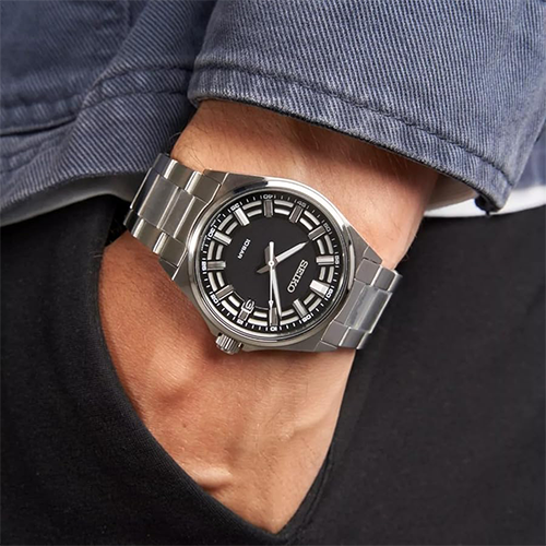 Silver watch with black face on a person's wrist, wearing a denim shirt and black jacket.